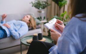 Unrecognizable professional therapist writing on clipboard while sitting against male client lying on couch on blurred background during mental health therapy - Photo by SHVETS production on Pexels