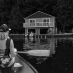 Woman paddling a canoe towards a lakeside house, capturing summer relaxation. - Photo by Paige Thompson on Pexels